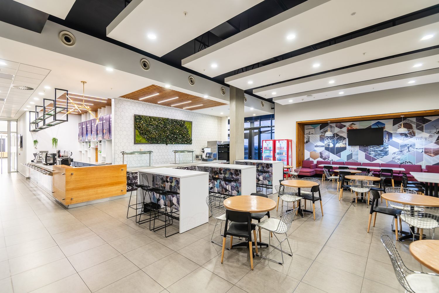 Modern cafe interior with bar seating, dining tables, green wall feature, and TV screens in a bright open space.
