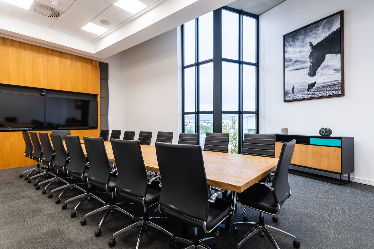 Modern conference room with large wooden table, black ergonomic chairs, floor-to-ceiling windows, and wall-mounted TV screen.