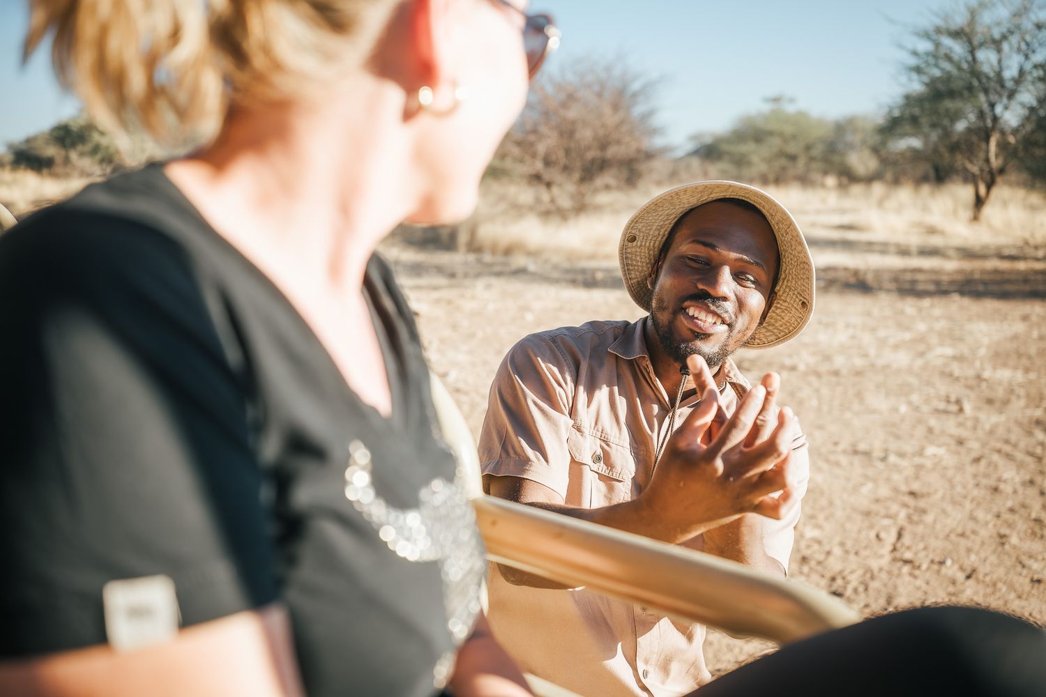 A smiling man in a straw hat converses with a woman outdoors in an arid African landscape on a sunny day.