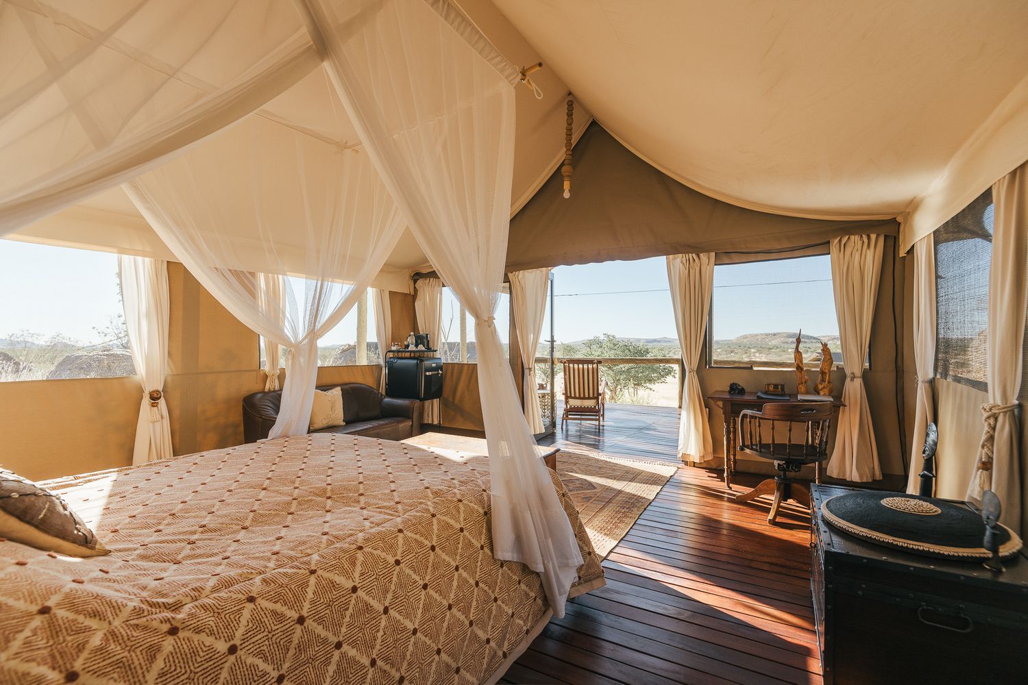 Luxury safari tent bedroom with canopy bed, sheer curtains, and open-air deck overlooking a scenic African landscape.