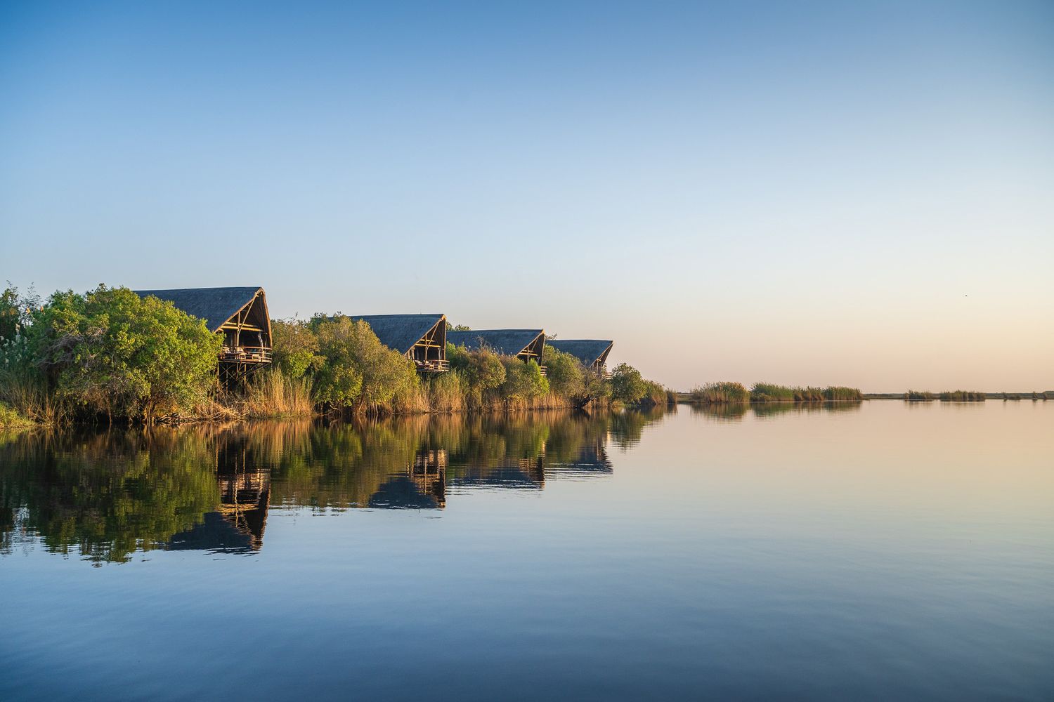 Luxury safari lodges reflecting in calm waters surrounded by lush vegetation at golden hour in Africa.
