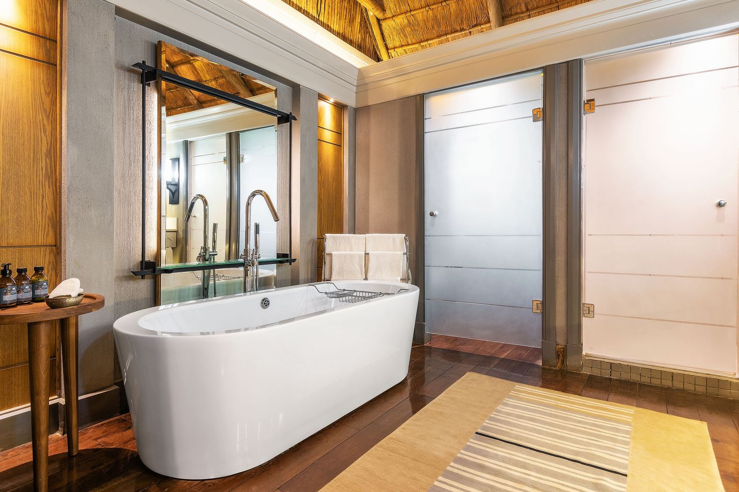 Elegant freestanding soaking tub in a luxury safari lodge bathroom with thatched roof and wood floors.