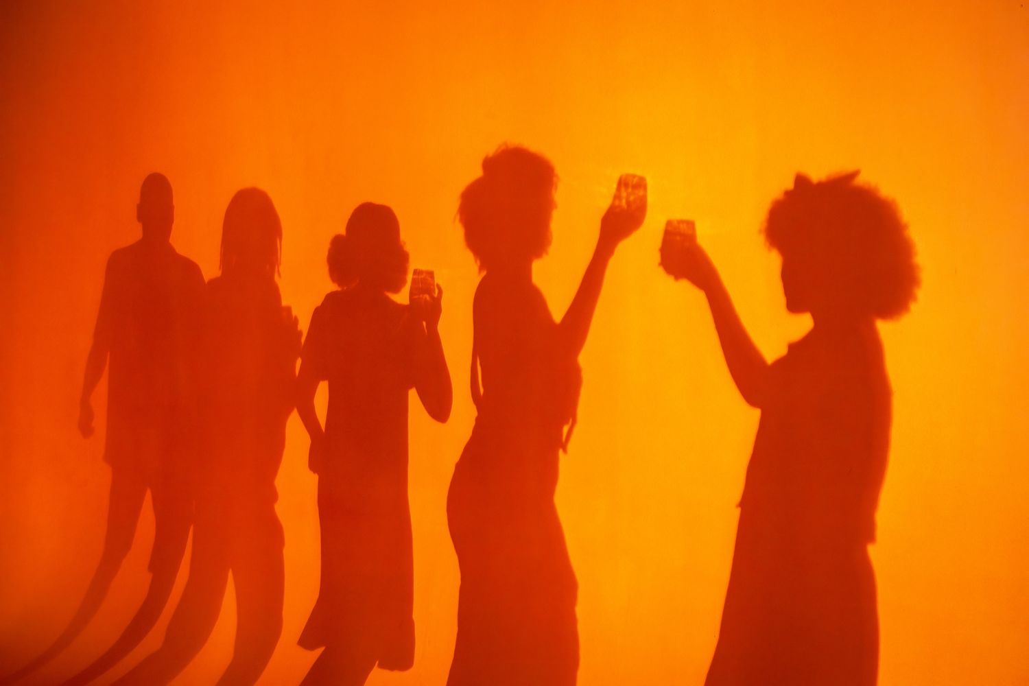 Silhouettes of people holding drinks cast against a vibrant orange background at a social gathering.