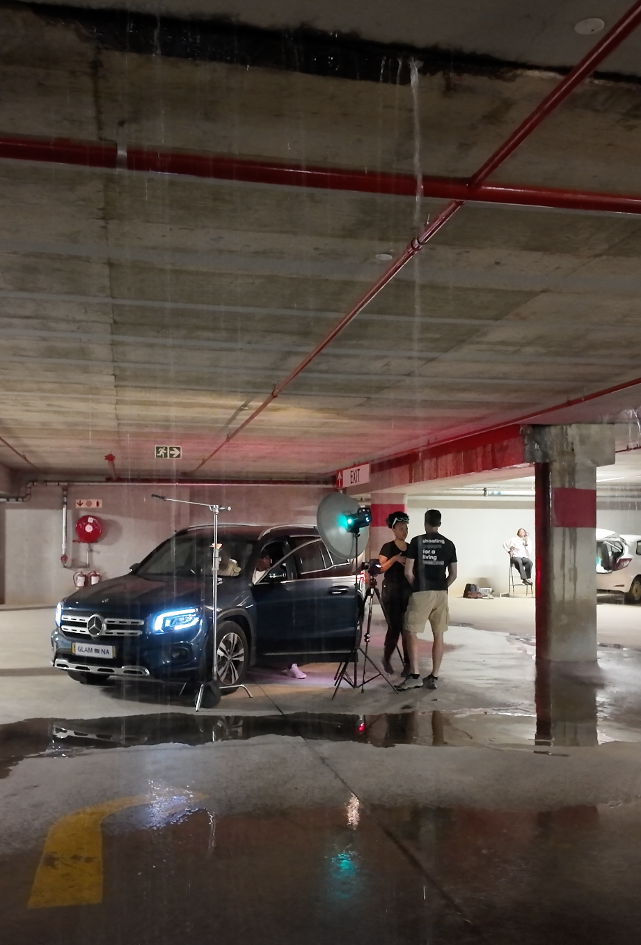 Two people filming a Mercedes-Benz SUV in a wet underground parking garage with dramatic lighting.