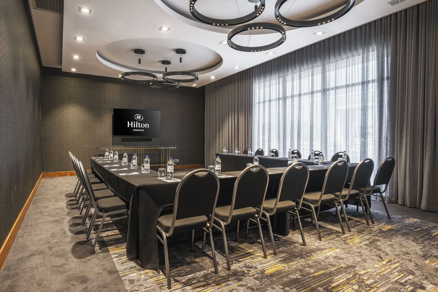 Hilton hotel conference room with U-shaped table setup, black chairs, circular ceiling lights, and a wall-mounted TV screen.