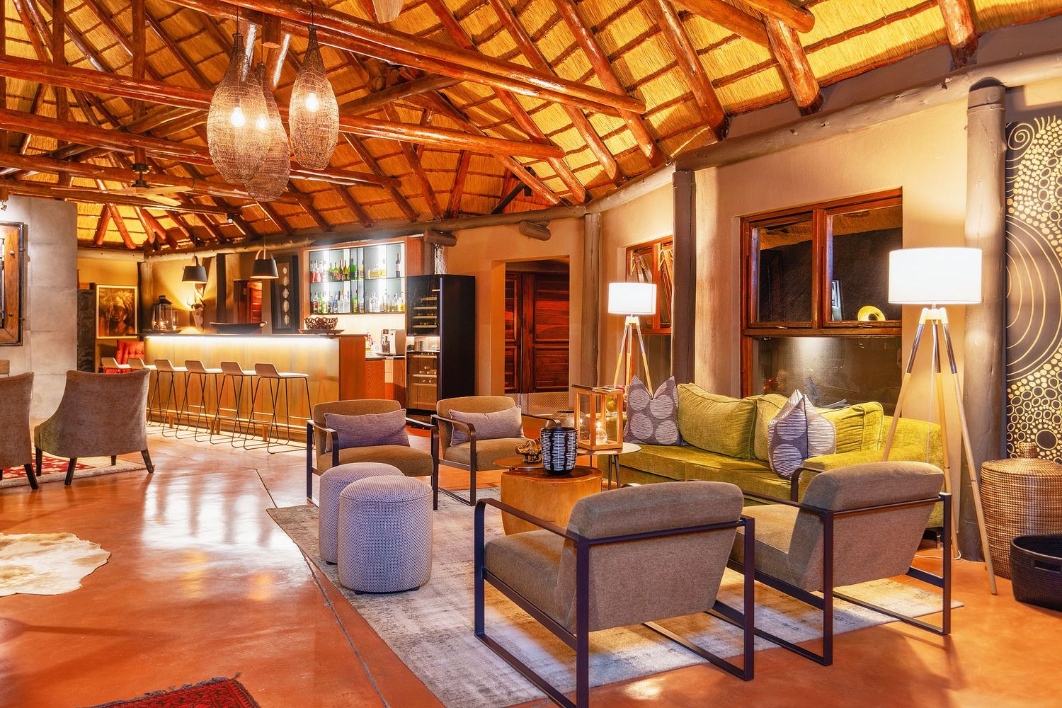 Warm safari lodge lounge with thatched ceiling, green sofas, and a well-stocked bar area.