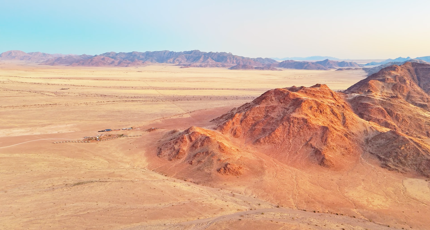 Aerial view of a vast desert landscape with rugged orange rock formations and distant mountains under a clear sky.