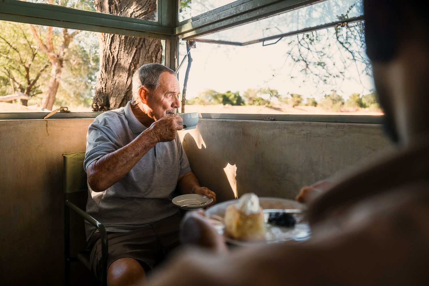 Older man drinking coffee at an outdoor cafe table in warm sunlight near a tree.