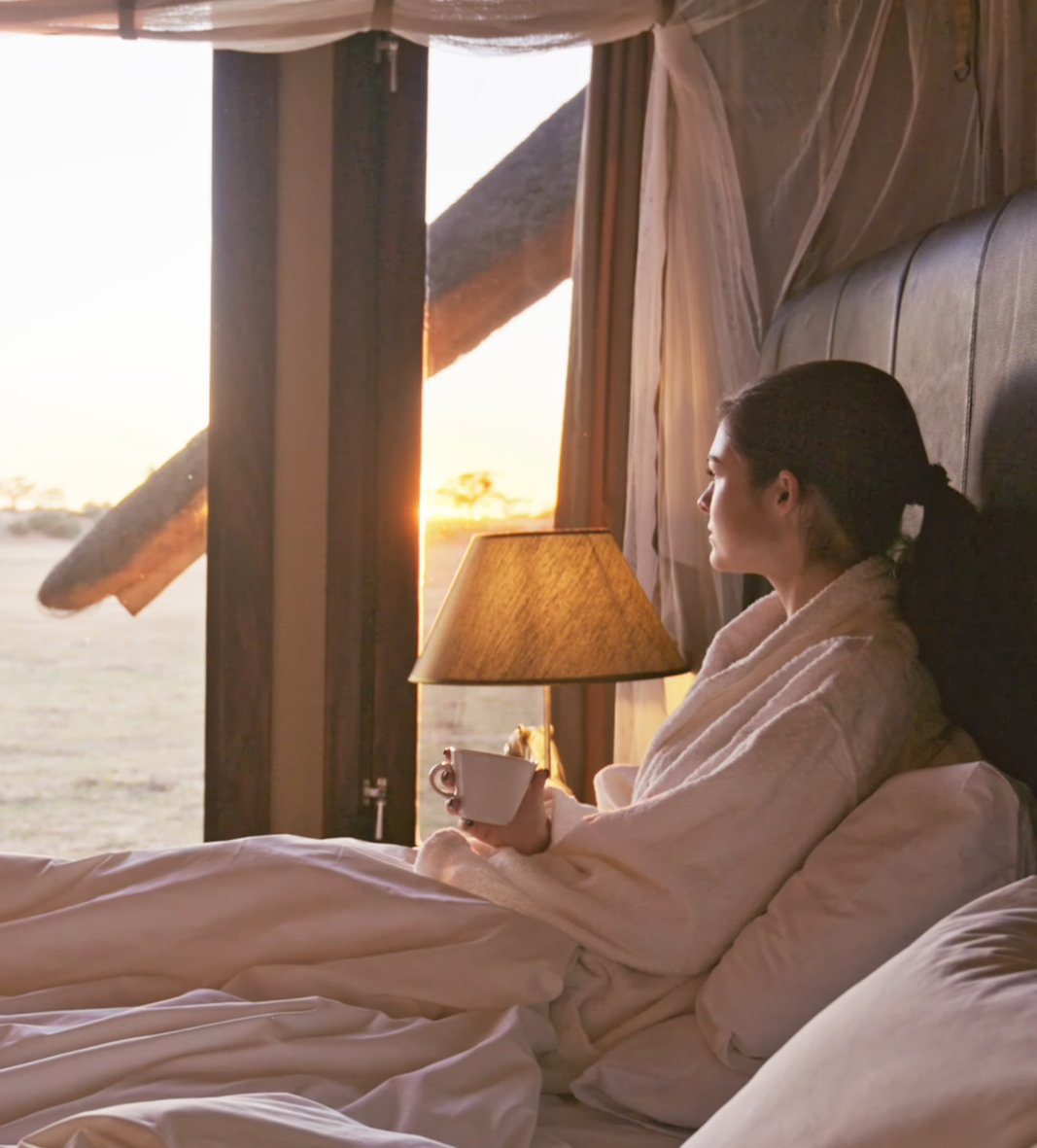 Woman in white robe sipping coffee in bed at luxury safari lodge, watching golden sunset through open canvas walls.
