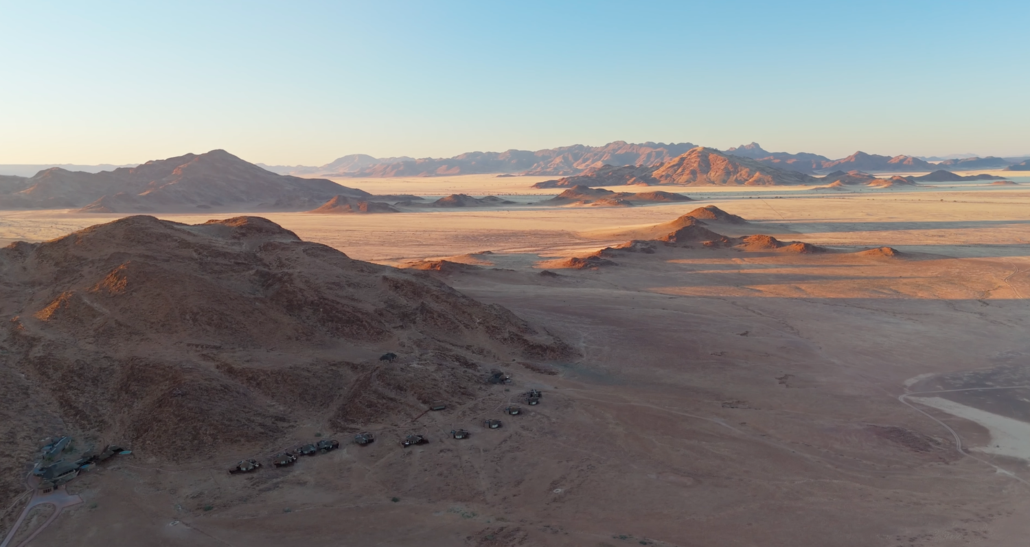 Aerial view of a vast arid desert landscape with rocky hills, sandy plains, and distant mountains at golden hour.