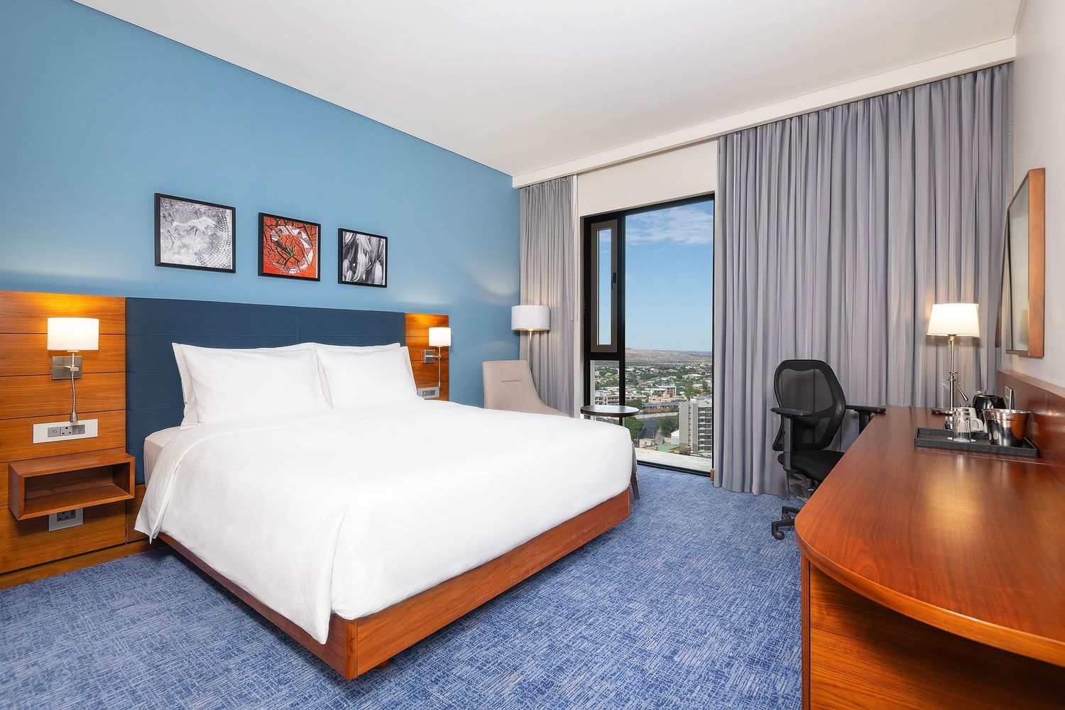 Modern hotel room with king bed, blue accent wall, city view window, and wooden desk on blue carpet.