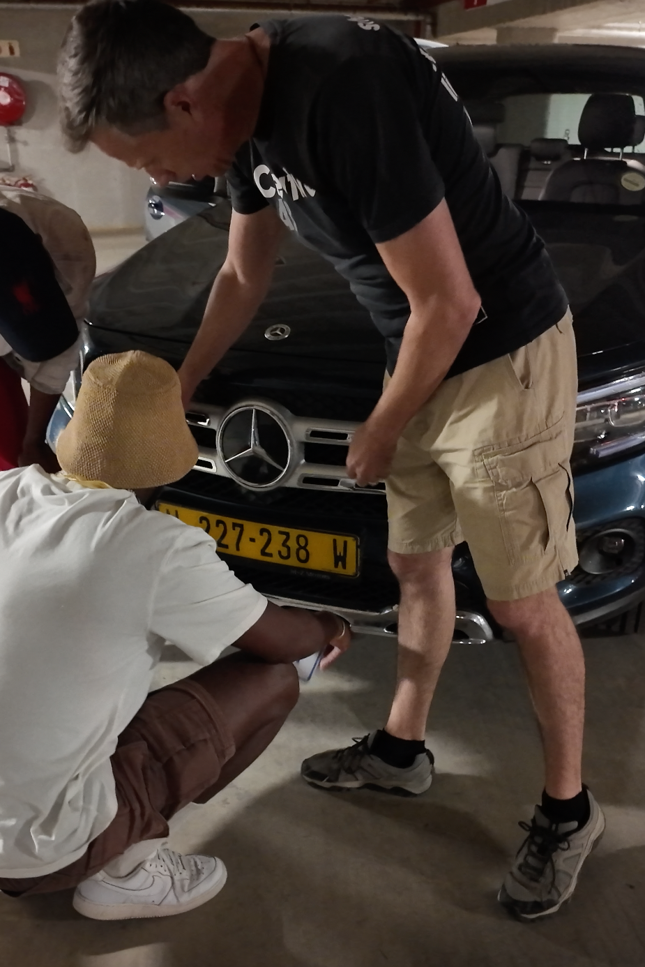 Two men inspecting the front bumper of a Mercedes-Benz car in a parking garage.