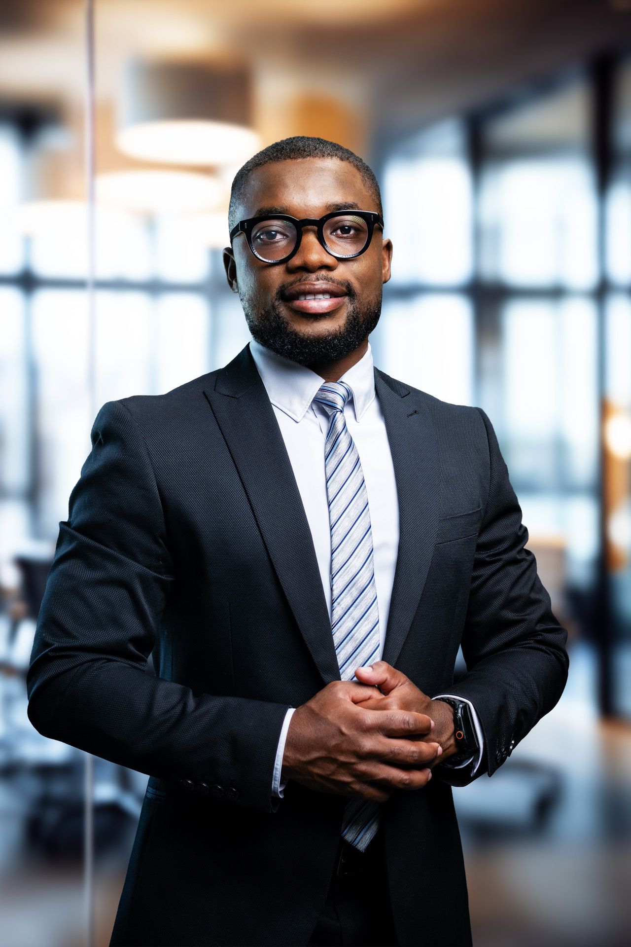 Professional Black businessman in dark suit and glasses stands confidently in modern office, hands clasped.