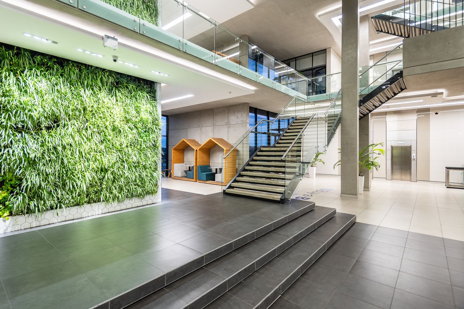 Modern office lobby with lush green living wall, concrete staircase, glass railings, and wooden pod seating areas.