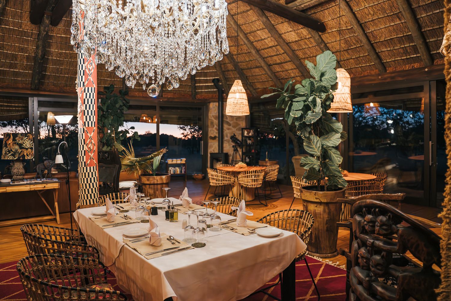 Elegant safari lodge dining room with crystal chandelier, white-clothed tables, rattan chairs, and tropical plants under thatched roof.