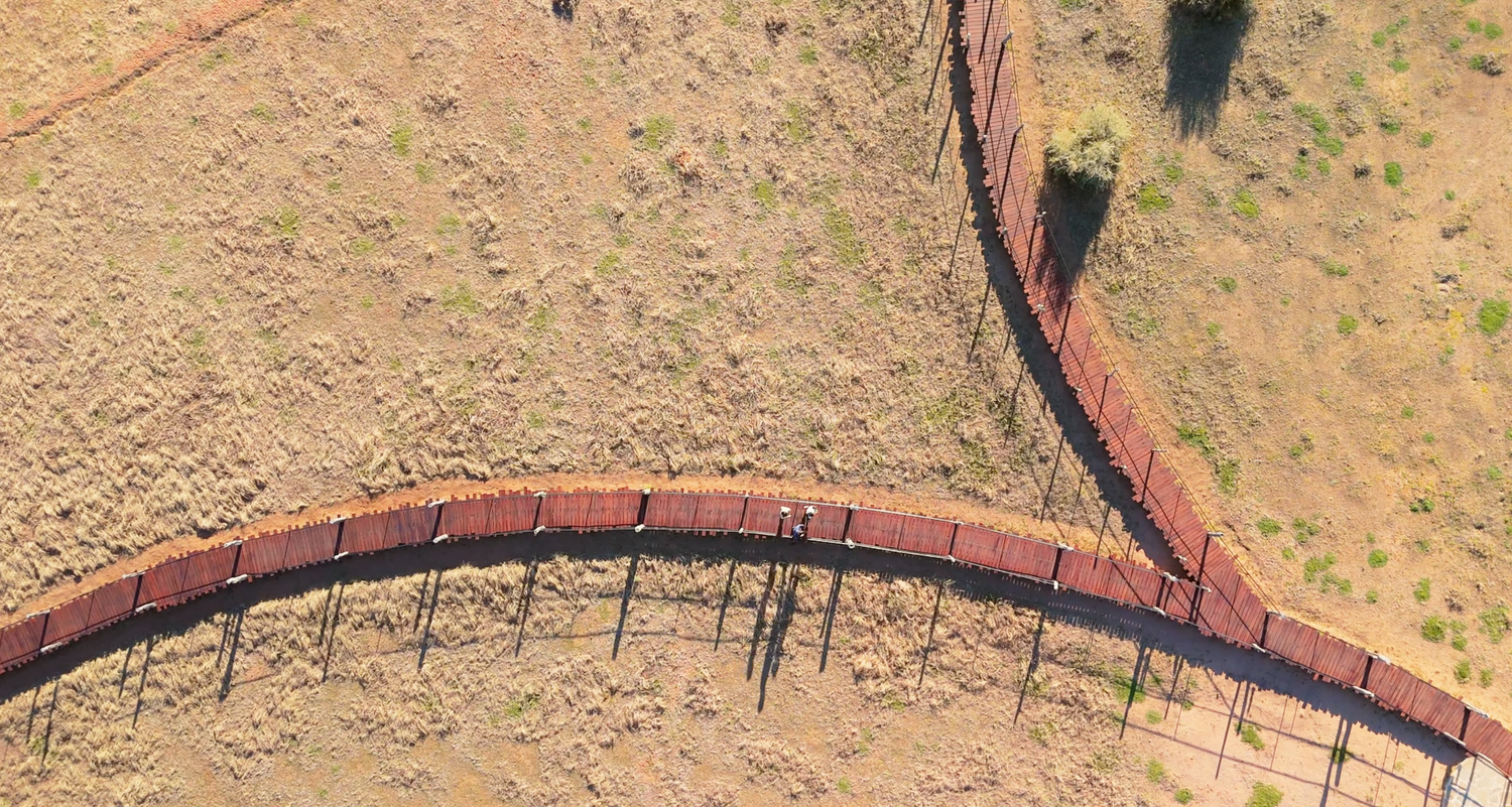 Aerial view of a curved rusty metal fence crossing dry arid terrain in outback Australia.