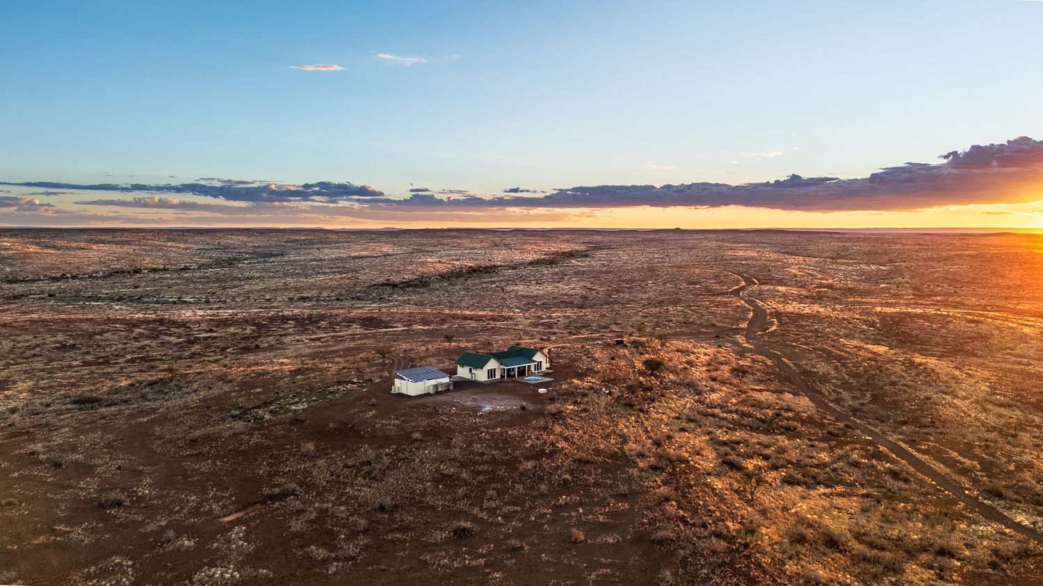 Aerial view of a small isolated farmhouse surrounded by vast flat arid plains under a vivid sunset.