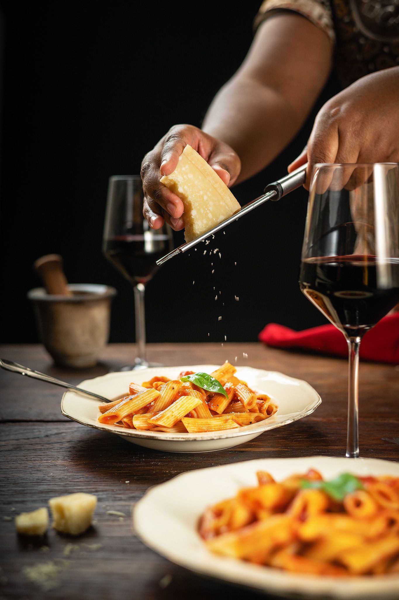 Chef grating fresh Parmesan cheese over penne pasta in tomato sauce, with red wine glasses on rustic wooden table.