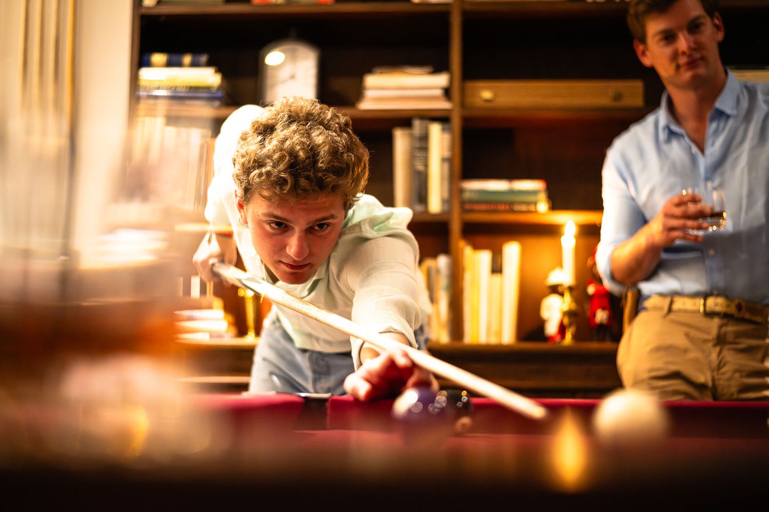 Young man leaning over a pool table taking a shot, with a friend watching in a warmly lit game room.