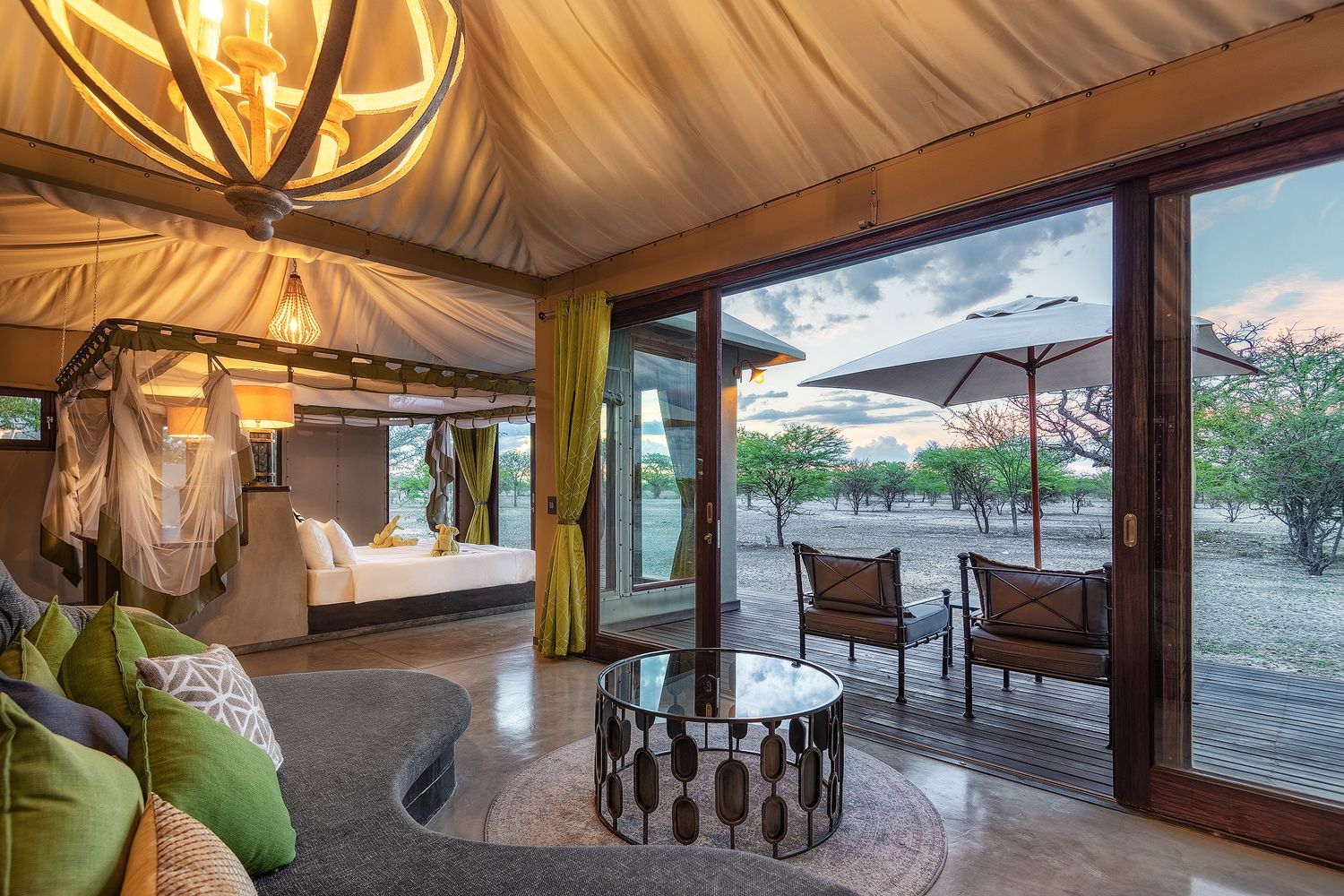 Luxury safari tent interior with canopy bed, green accents, and open glass doors revealing an outdoor seating area at dusk.