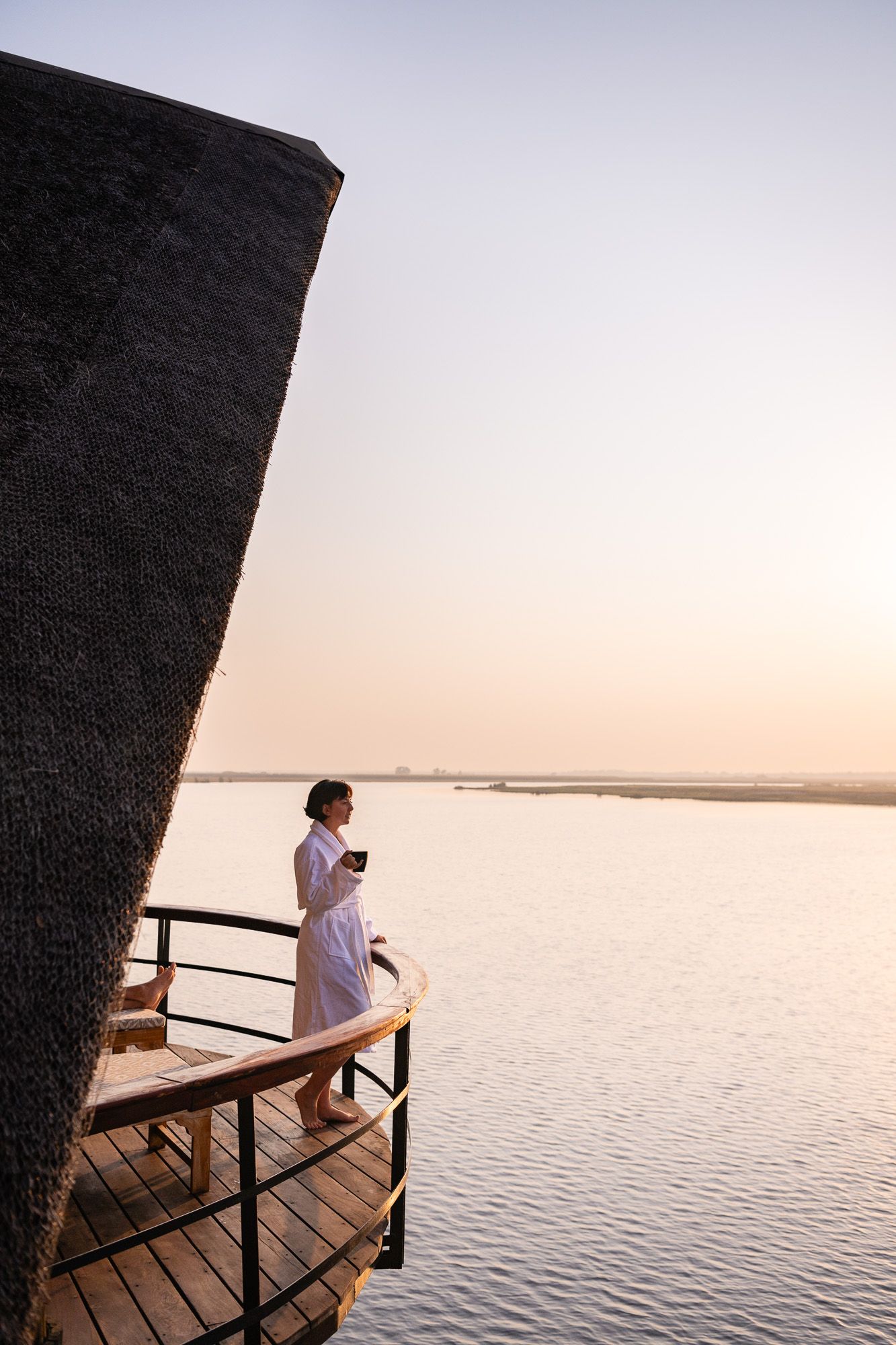 Woman in white robe holding coffee cup on wooden deck overlooking calm river at golden sunset.
