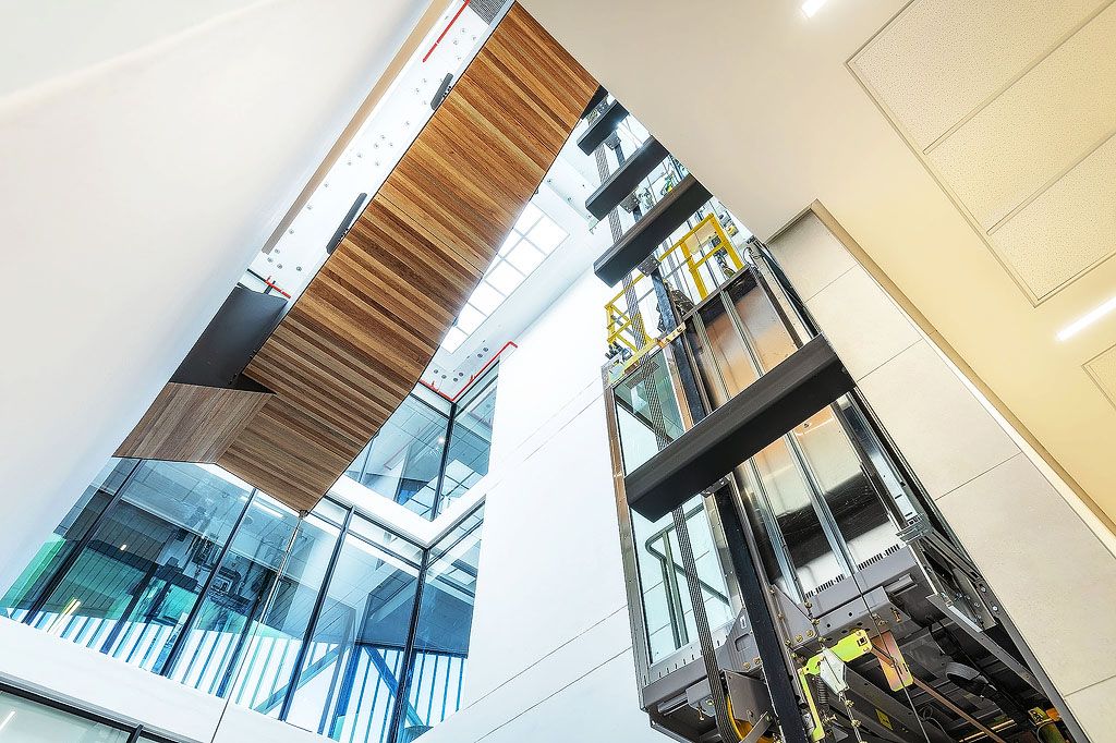 Striking upward perspective of modern office atrium with wooden ceiling feature, glass elevator shaft, and natural light.