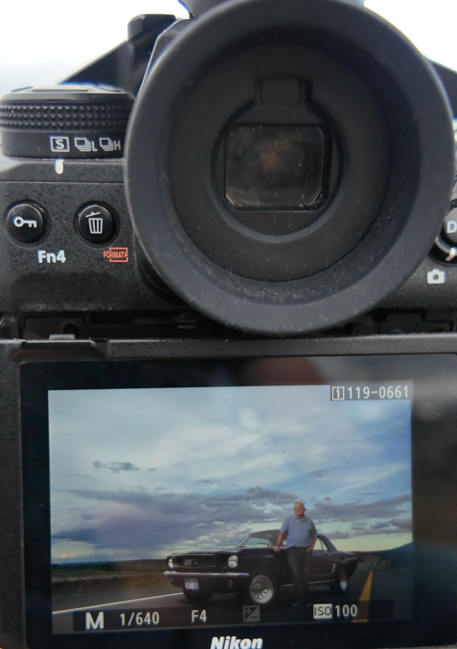 Close-up of a Nikon camera LCD screen displaying a man posing beside a classic car under a dramatic cloudy sky.