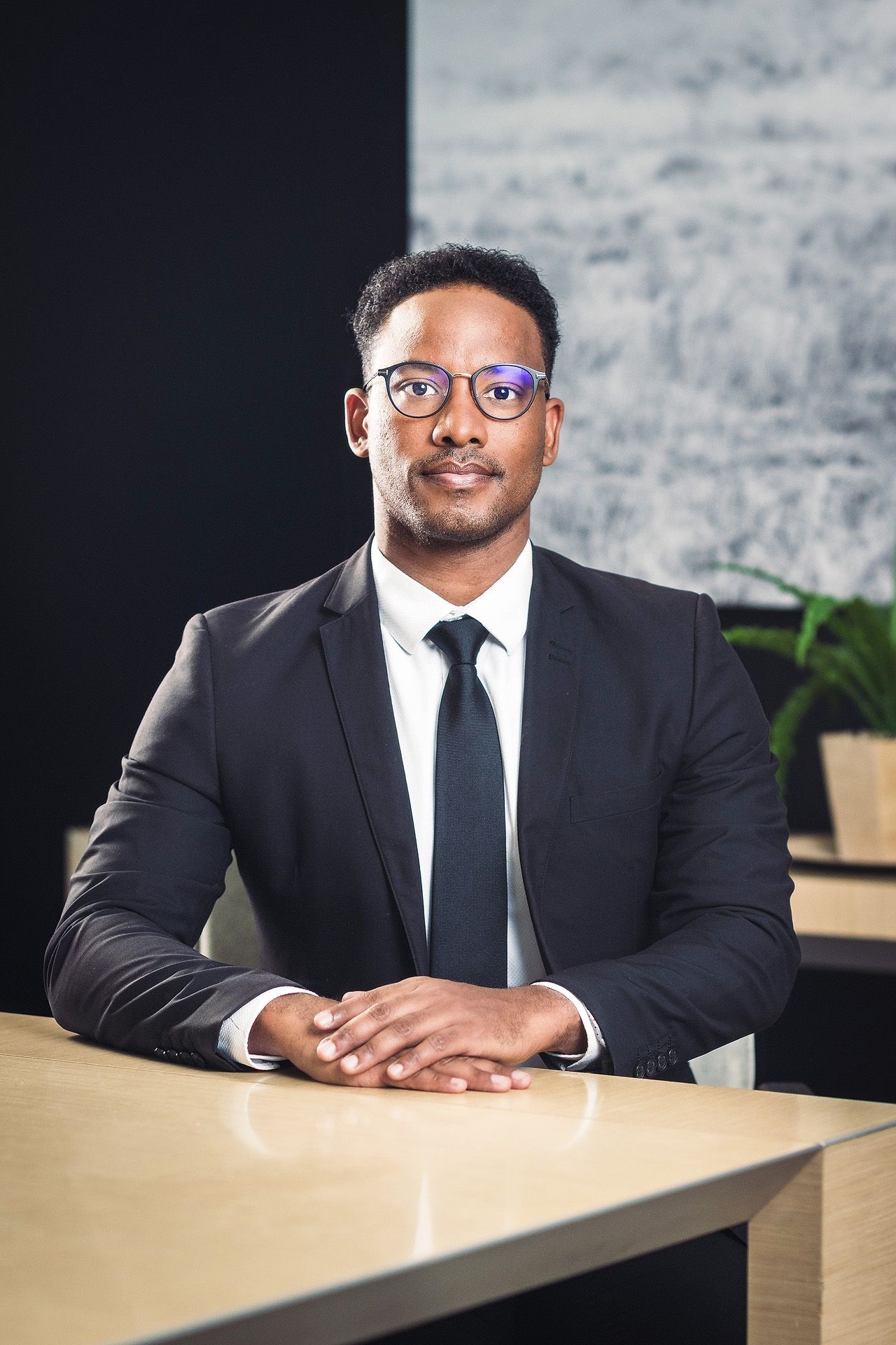 A professional Black man in a dark suit and tie with glasses sits confidently at a wooden desk in a modern office.