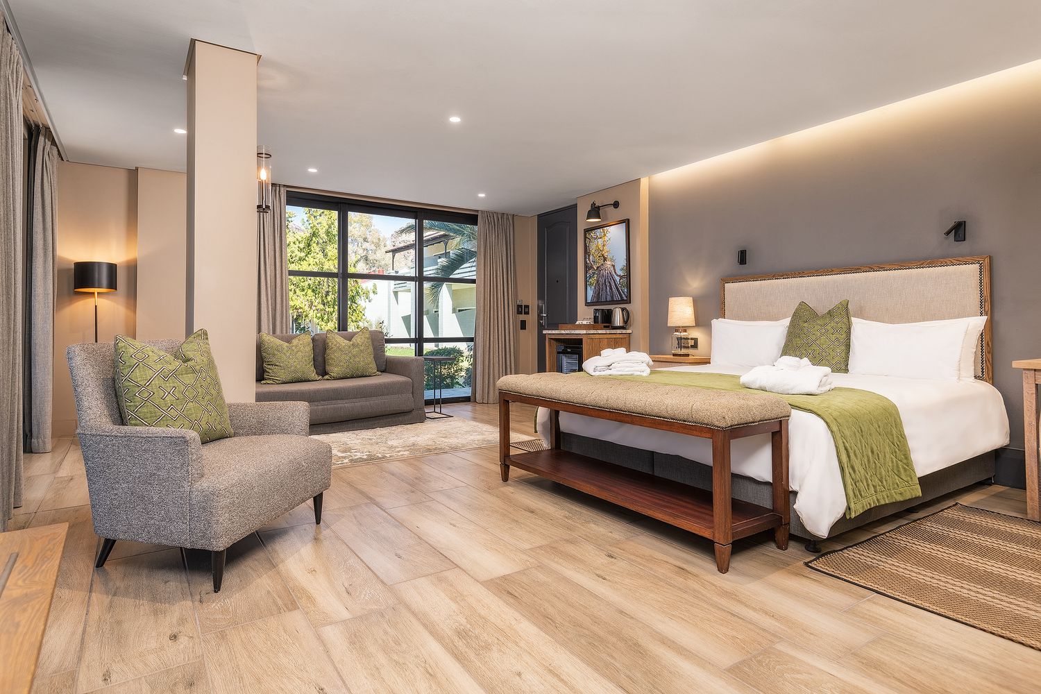 Elegant hotel suite with hardwood floors, king bed, green accents, gray armchair, and floor-to-ceiling windows overlooking greenery.