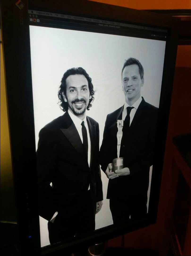 Two men in black suits posing with an award trophy in a black and white photo displayed on a monitor.