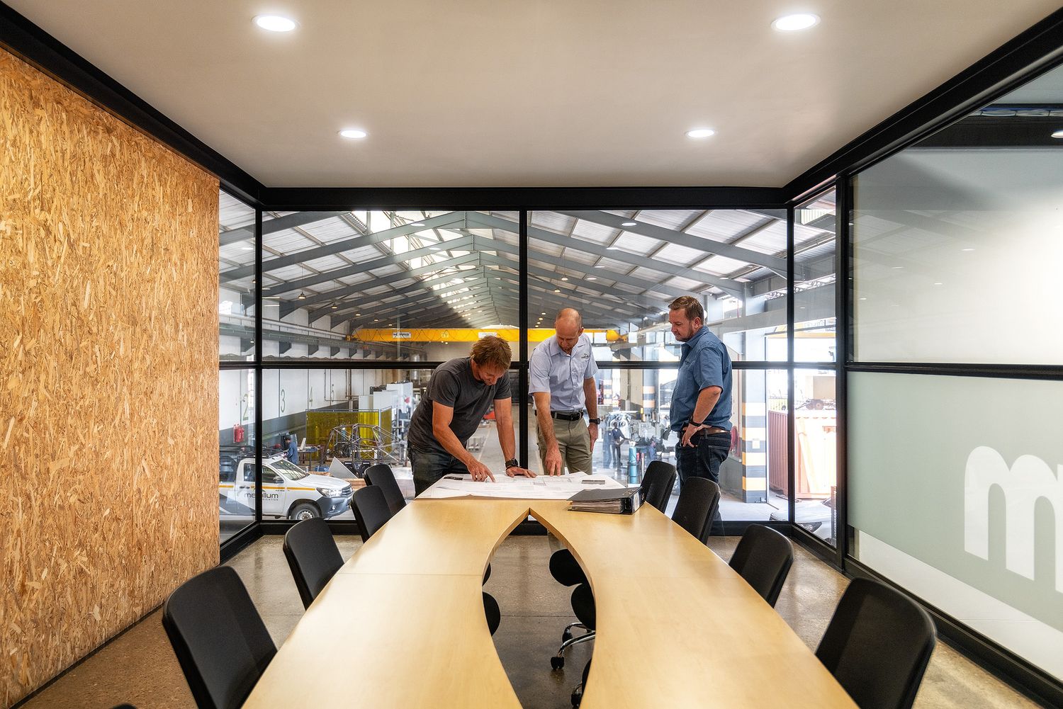 Three professionals reviewing blueprints in a modern conference room with floor-to-ceiling windows overlooking an industrial workspace.