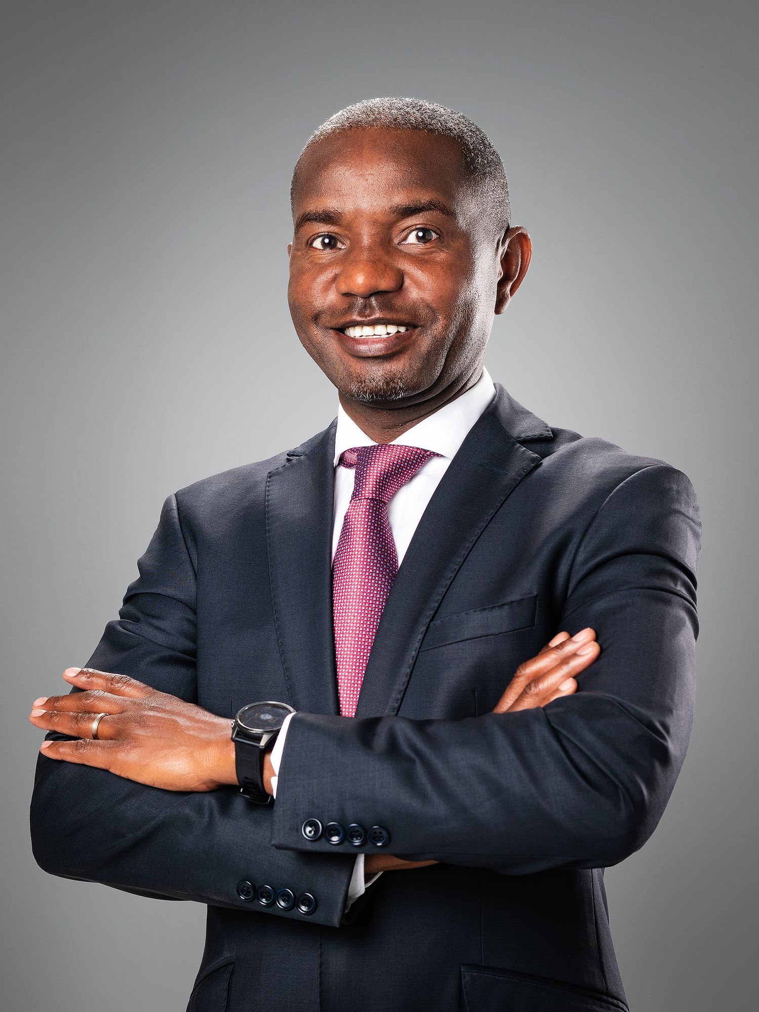 Professional businessman in dark suit and pink tie posing confidently with arms crossed against gray background.