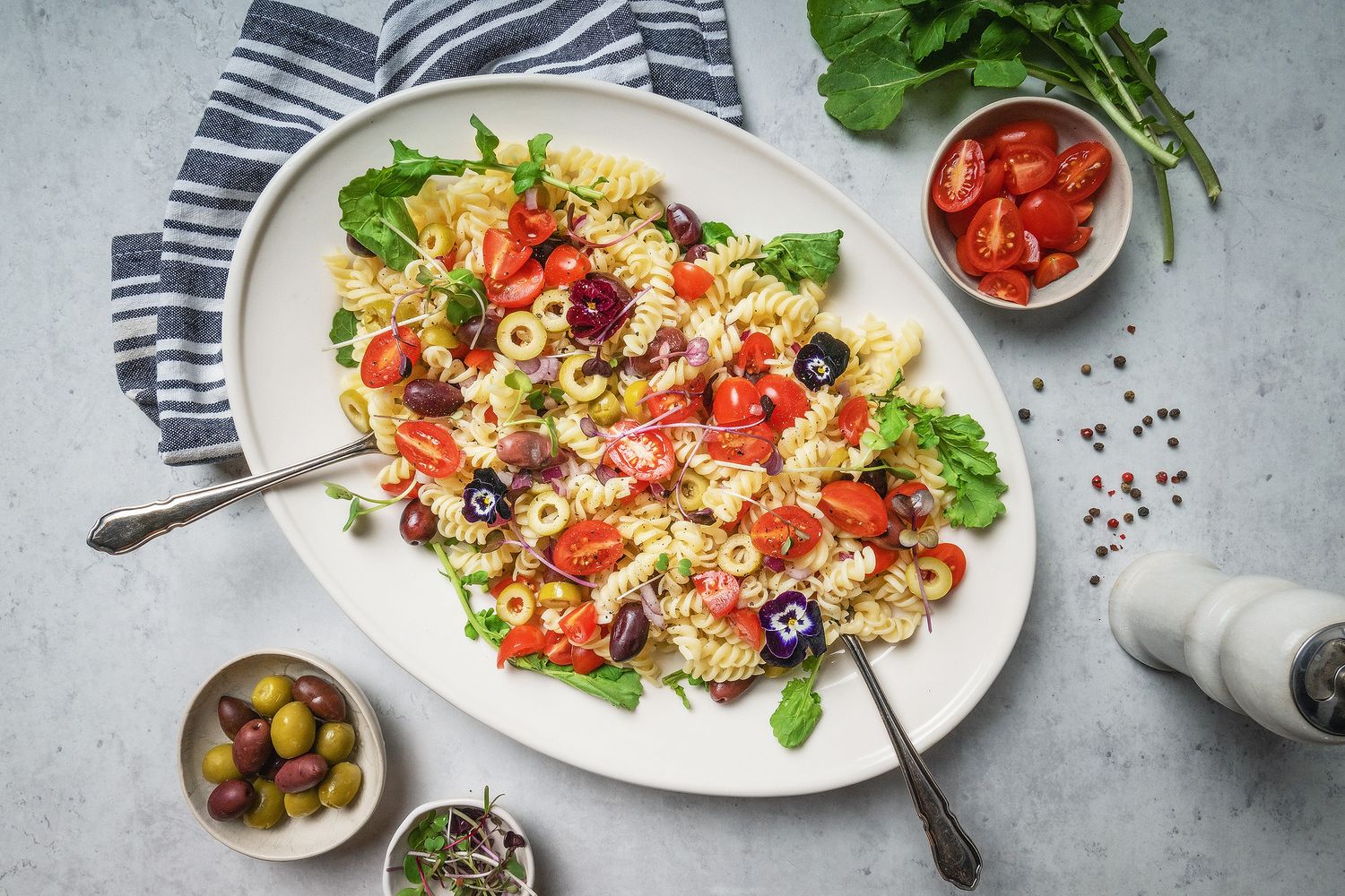 Overhead view of a white platter of rotini pasta salad with tomatoes, olives, and fresh herbs on a light surface.