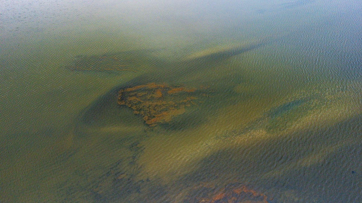 Aerial view of shallow lake with submerged rust-colored algae formations.