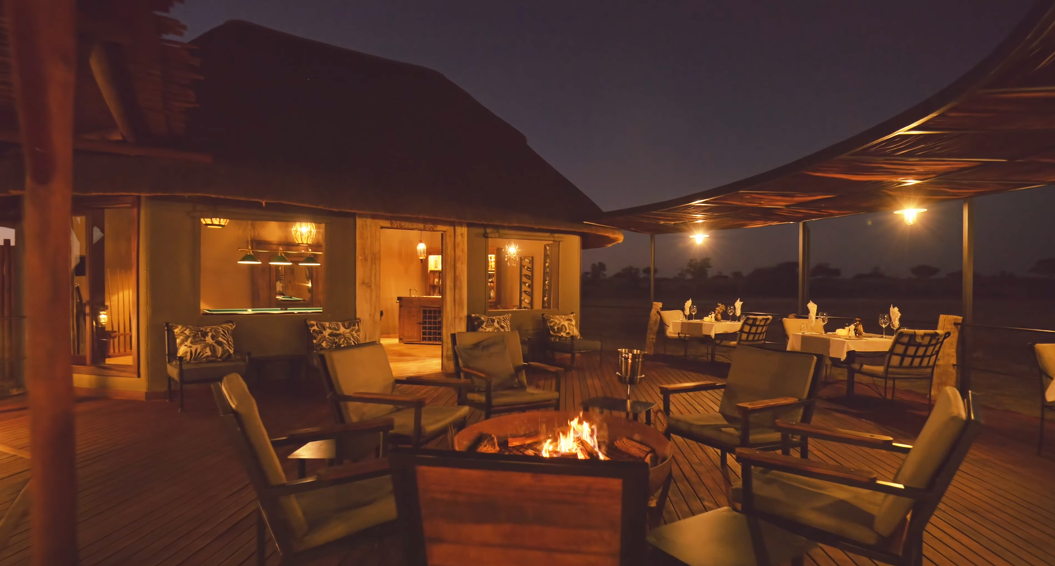 Elegant safari lodge terrace with firepit, plush seating, and warm ambient lighting at dusk overlooking the savanna.