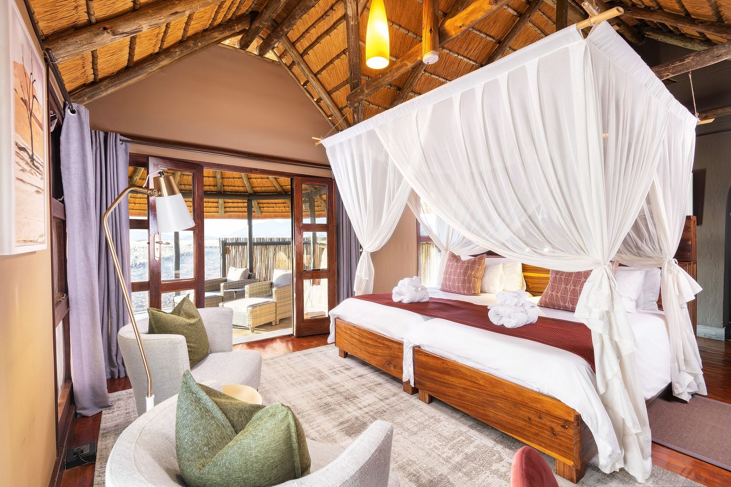 Elegant safari lodge bedroom with canopy bed, white drapes, wooden furniture, and thatched ceiling.