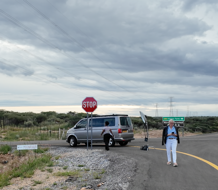 A man stands beside a silver van at a rural stop sign intersection under cloudy skies in an arid landscape.