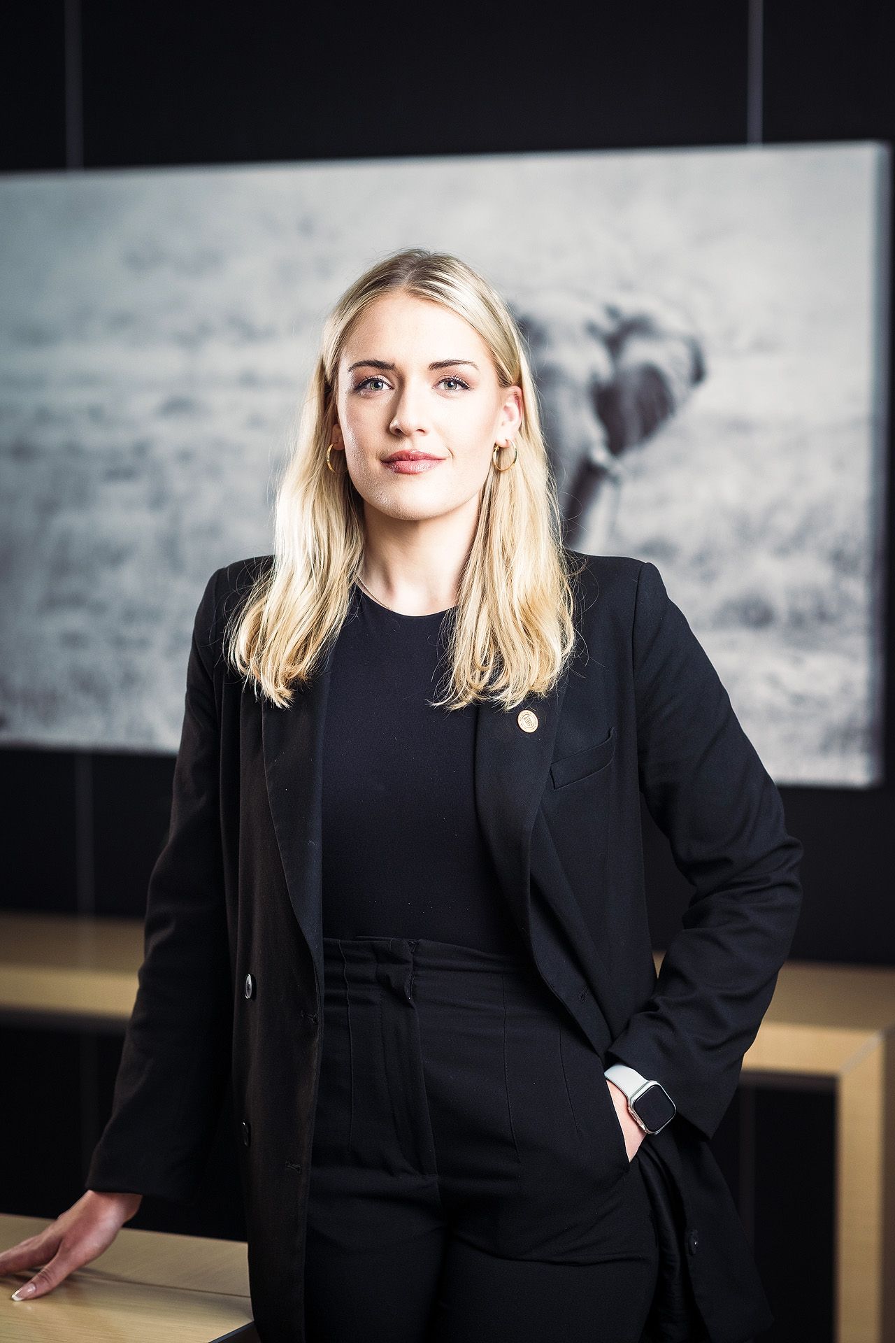 Professional blonde woman in black blazer and pants poses confidently in modern office with wildlife art.