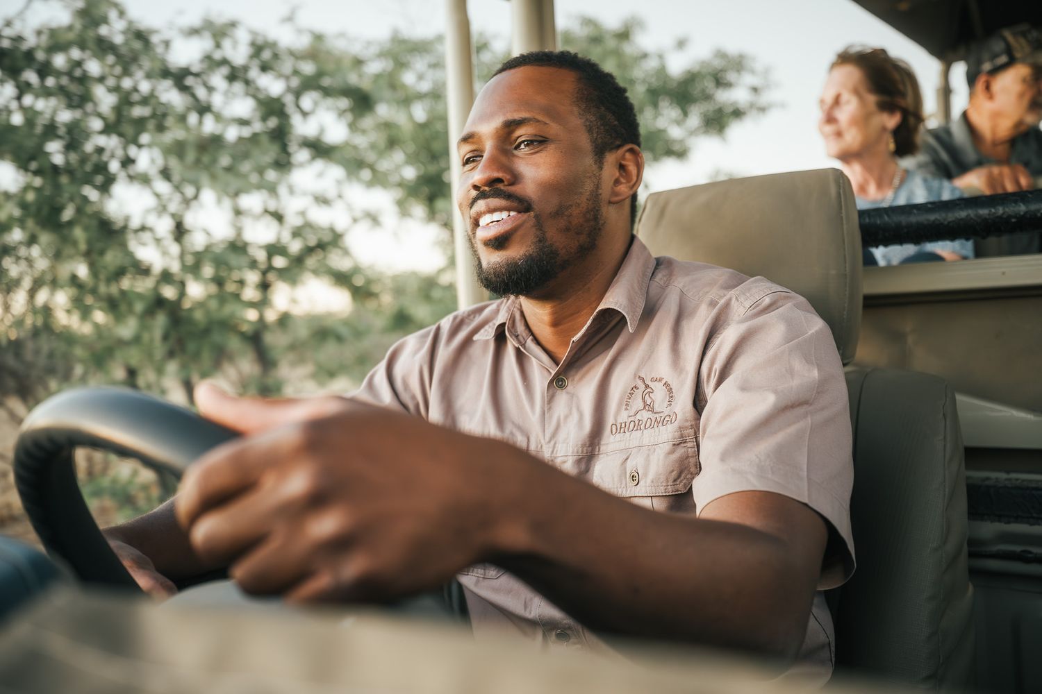 A smiling safari guide drives a game vehicle with tourists seated behind him in an open-air African bush setting.