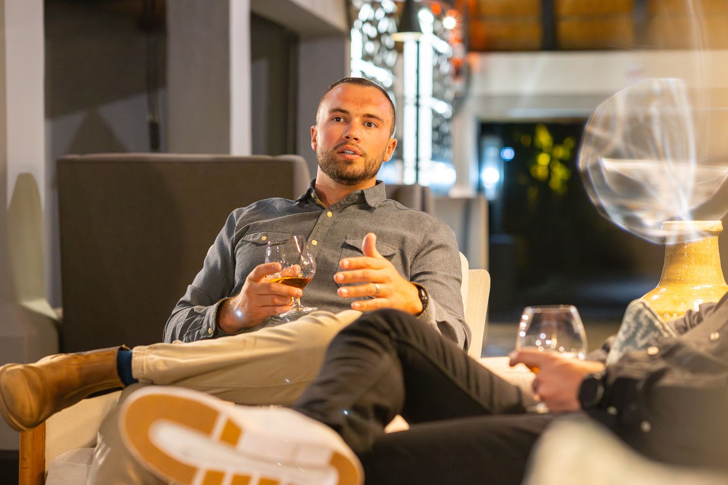 Man relaxing in chair holding whiskey glass during evening social gathering indoors.
