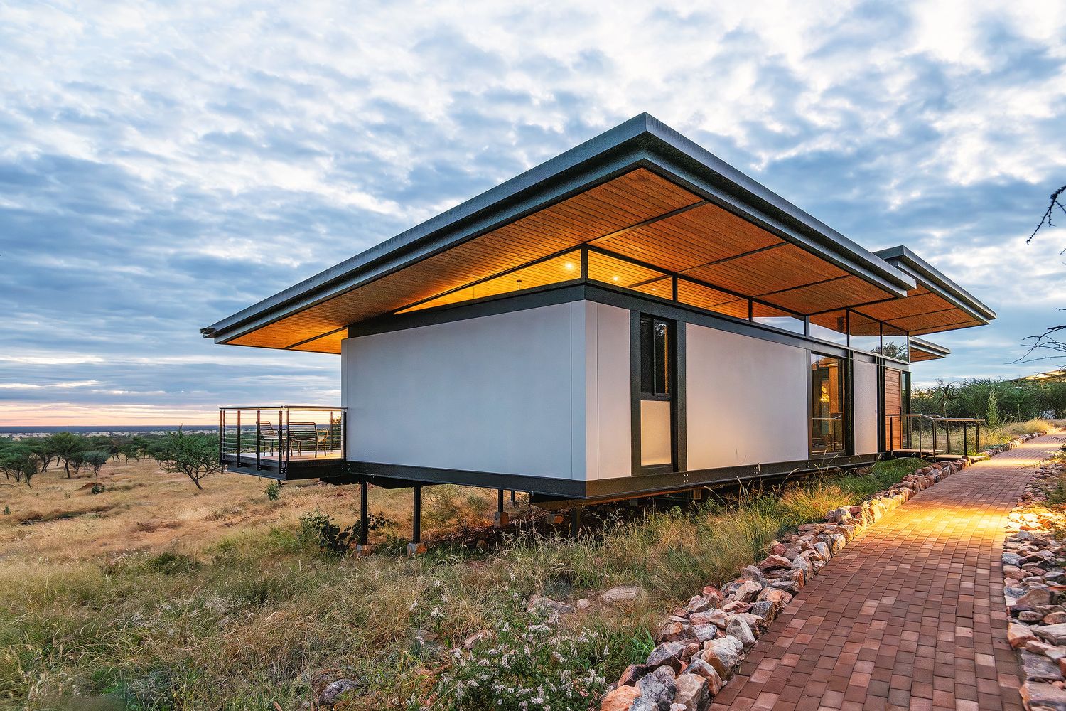 Modern elevated safari chalet at sunset surrounded by African bushveld with brick pathway leading up.