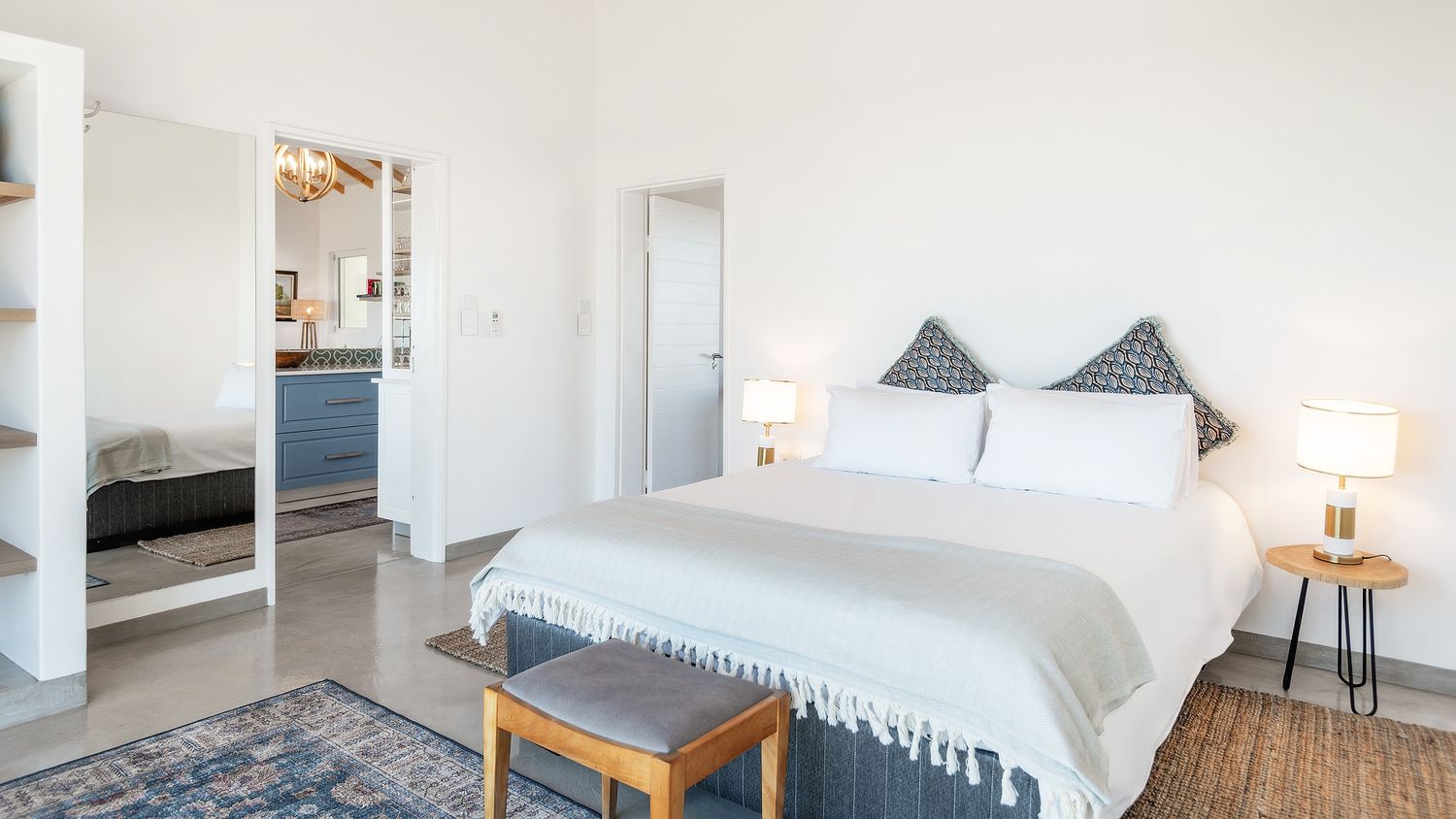 Bright white bedroom with a large bed, blue accent pillows, wooden stool, and an ensuite bathroom visible in the background.