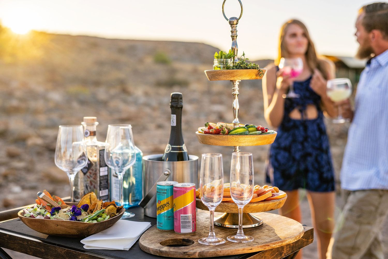 Outdoor sunset gathering with wine glasses, champagne, tiered appetizer stand, and couple enjoying drinks in desert setting.