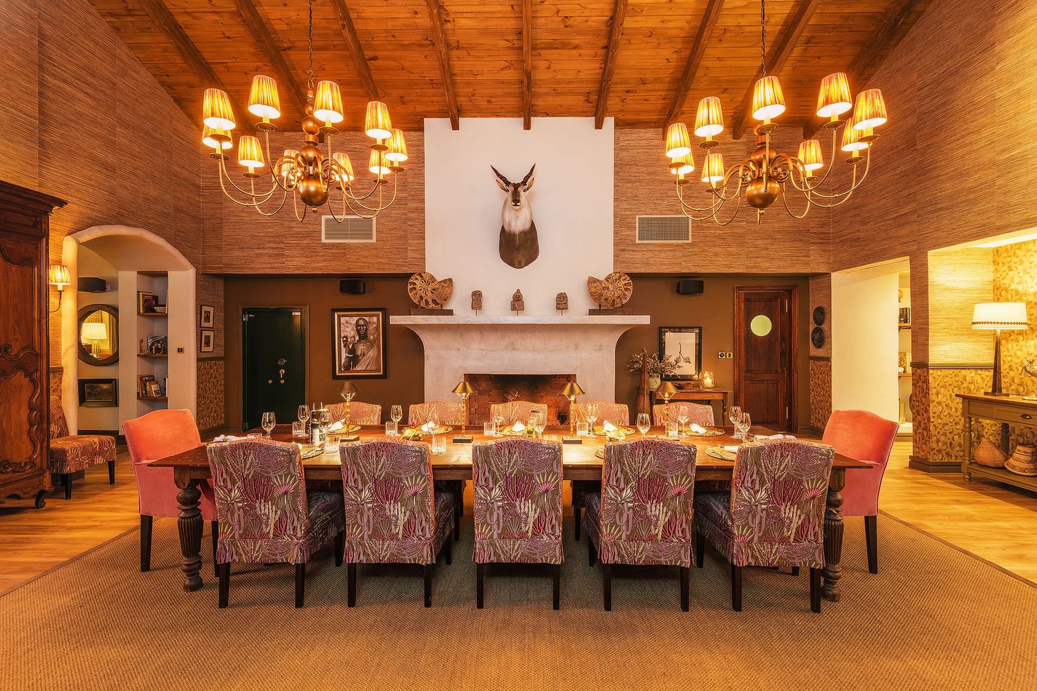 Elegant lodge dining room with long table, patterned chairs, fireplace, chandelier, and mounted animal trophy above mantle.