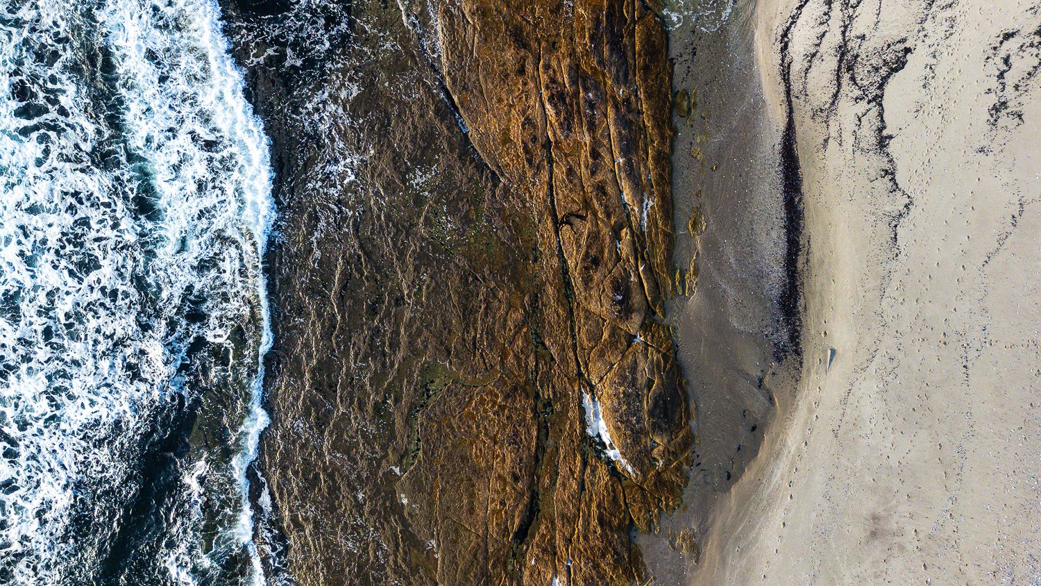 Aerial view of rocky coastline with crashing ocean waves meeting a sandy beach shore.