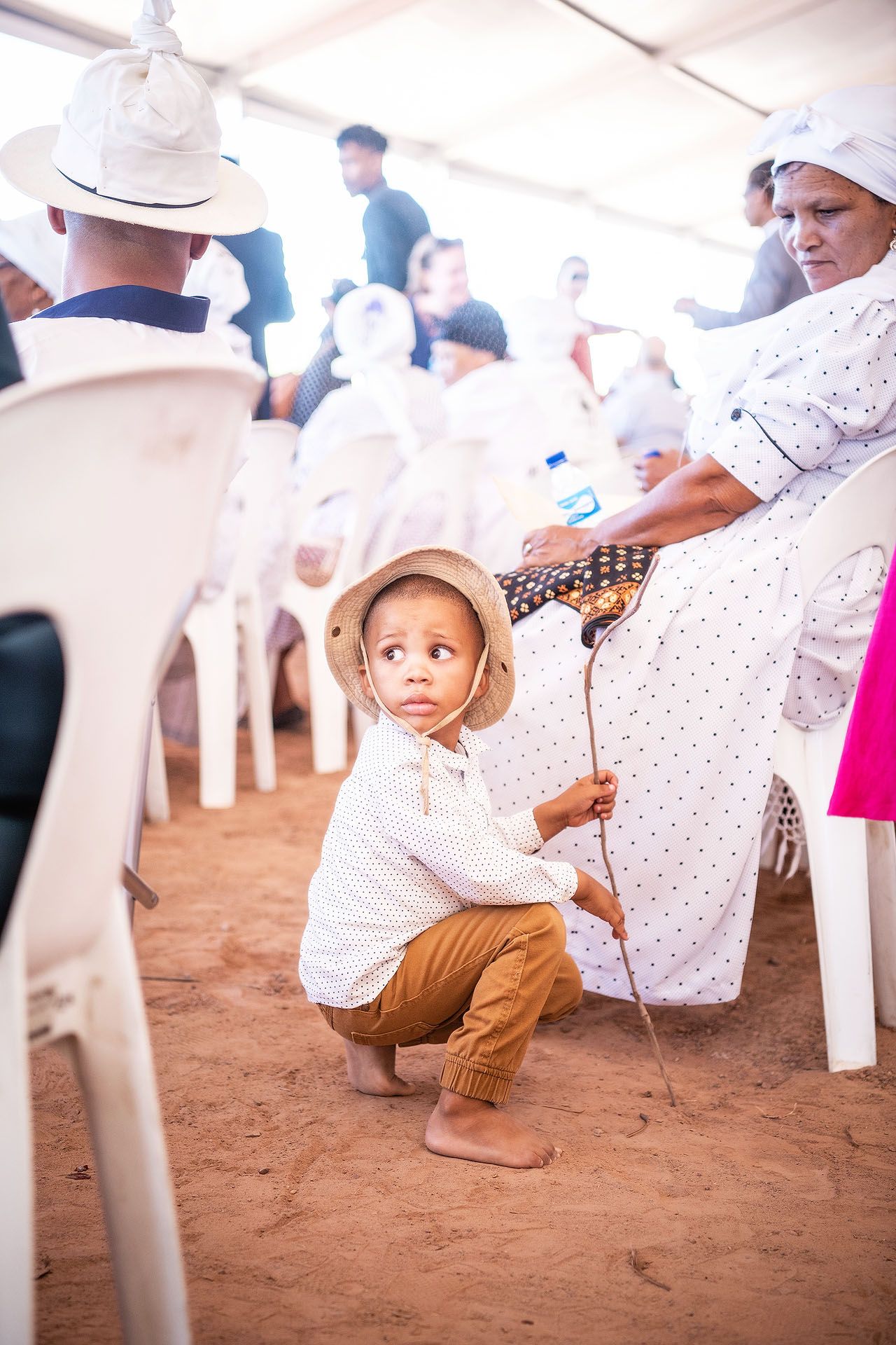 Young child in straw hat crouching barefoot on red soil during an outdoor gathering with adults seated on white chairs.