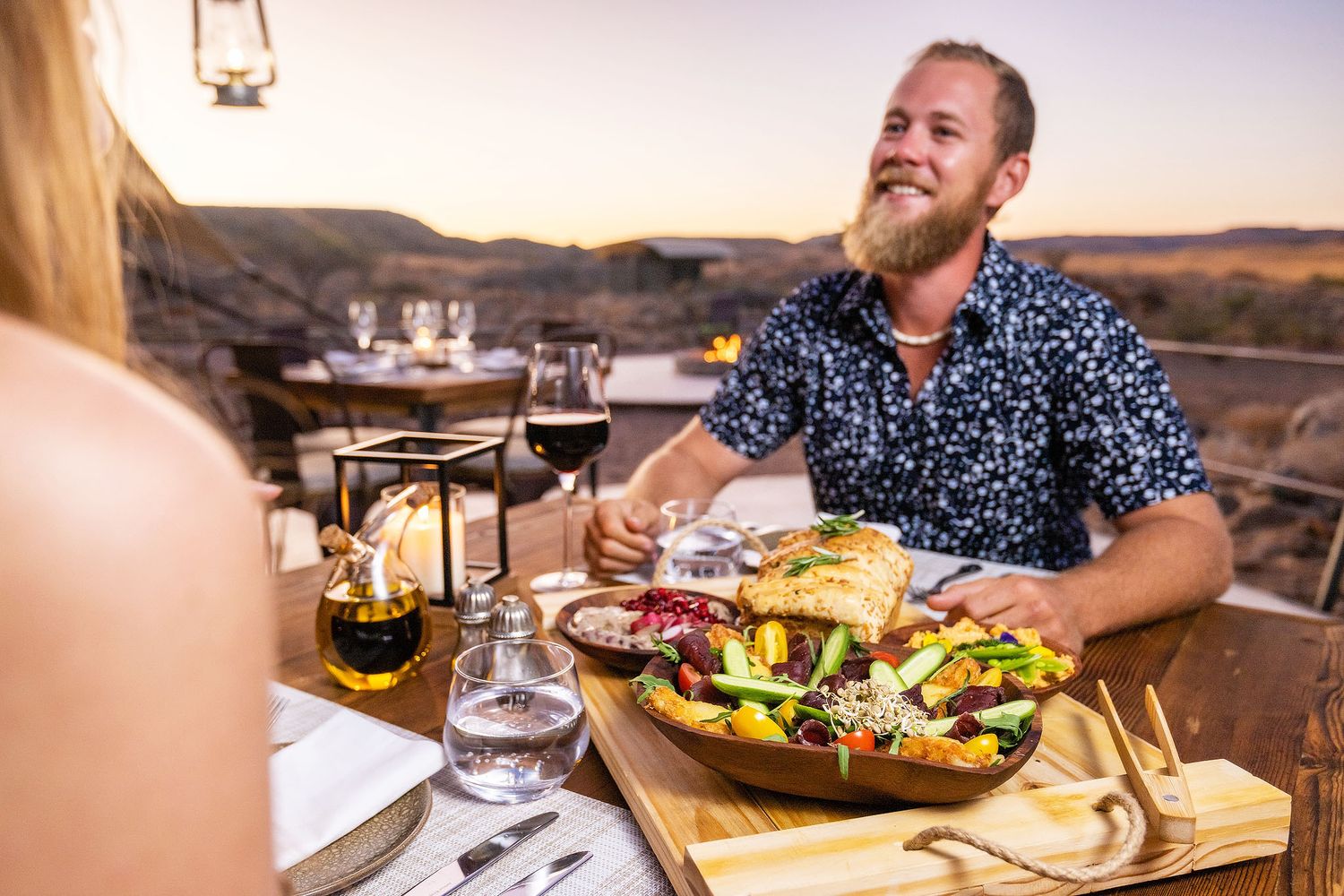 Man with beard dining outdoors at sunset, enjoying a colorful salad bowl with red wine in a desert landscape.