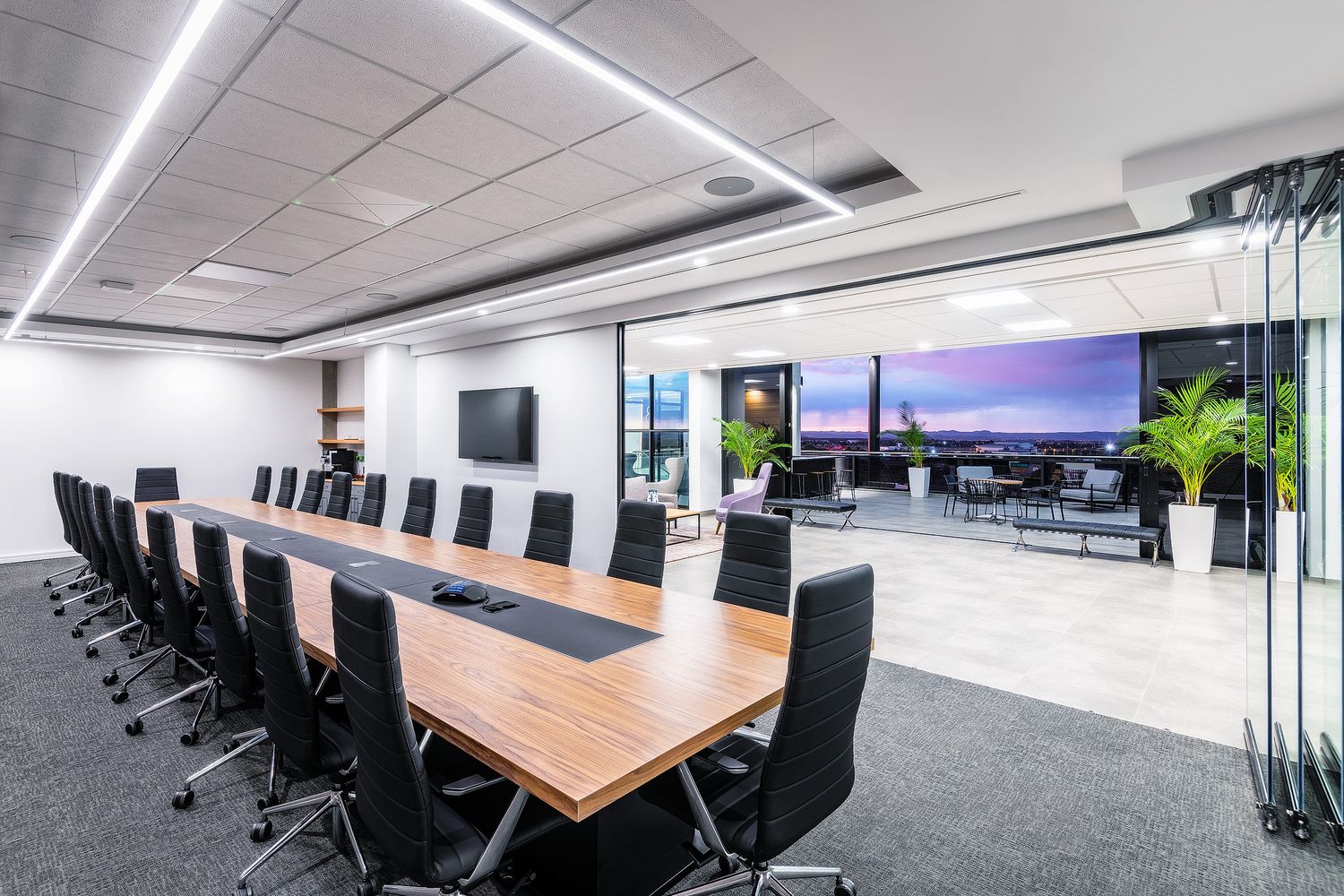 Sleek corporate boardroom with long wooden table, black chairs, wall-mounted TV, and floor-to-ceiling city view windows.