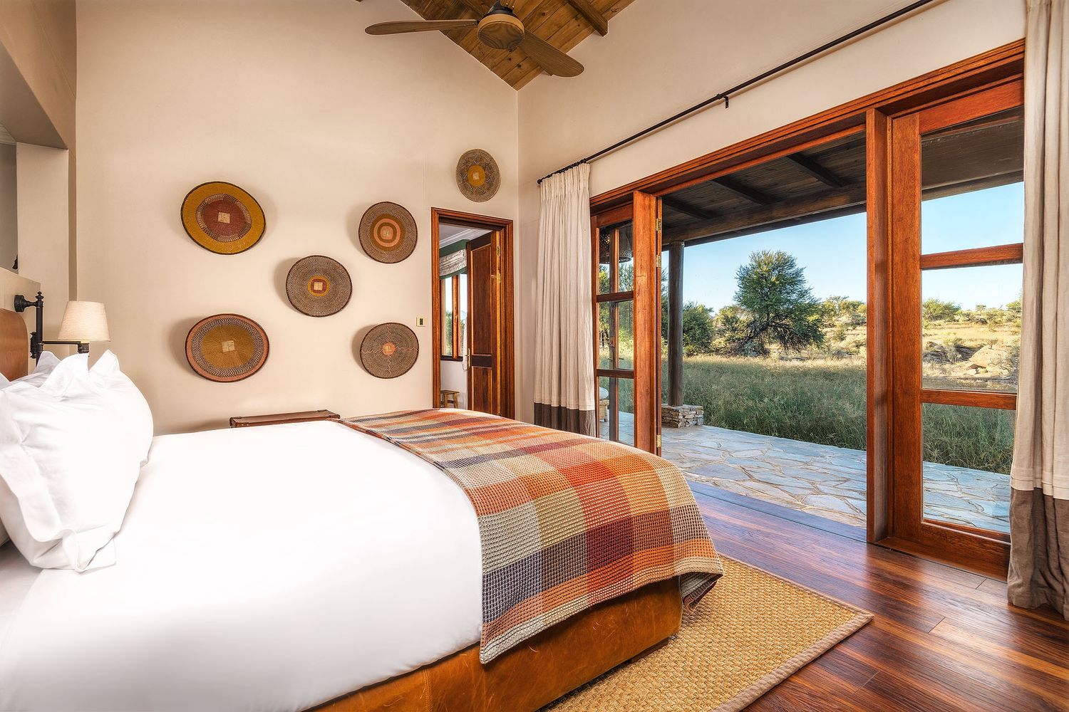 Elegant safari lodge bedroom with wooden floors, woven wall decor, plaid bedding, and glass doors opening to a scenic savanna view.