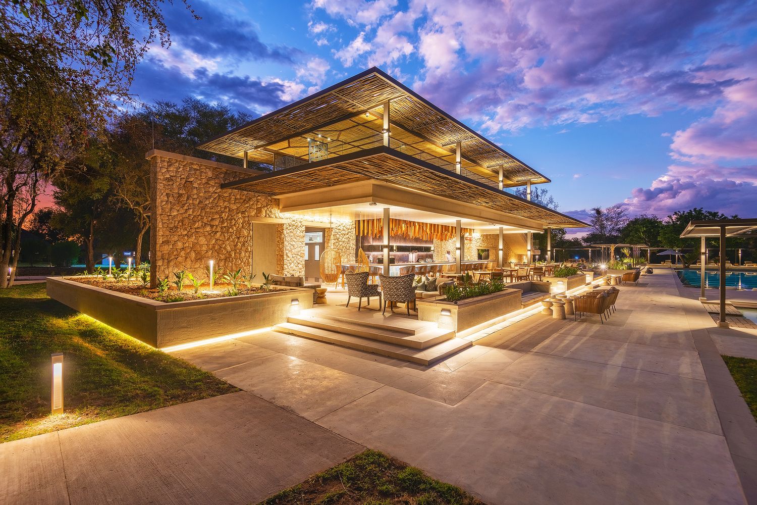Luxury resort pavilion illuminated at dusk with warm lights, stone walls, open-air dining, and a pool surrounded by lush gardens.